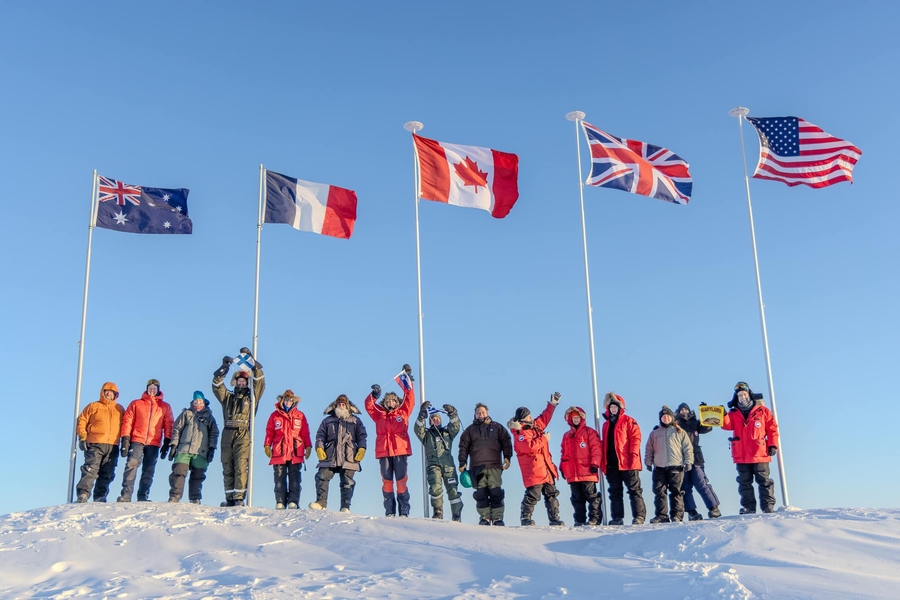 International team of approximately 20 people in winter gear standing on snow beneath flags of Australia, France, Canada, United Kingdom, and United States.