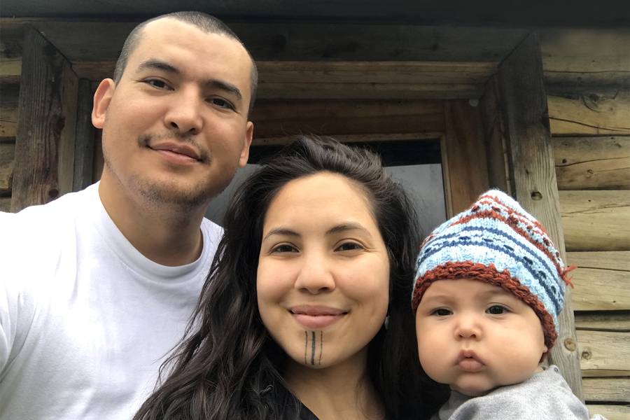 Family of three—man, woman with traditional facial tattoos, and young child in knit beanie—posed in front of log cabin structure.