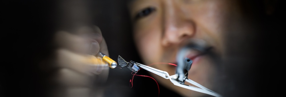 A close-up image of a person working with small electronic components. The person is holding a yellow-handled tool and manipulating wires and a small circuit board held by a white clamp.