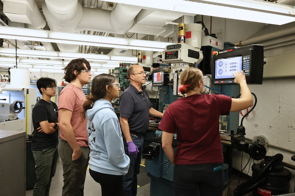 A diverse group of five people, wearing safety glasses in a workshop, listen to an instructor explaining a machine's digital interface.