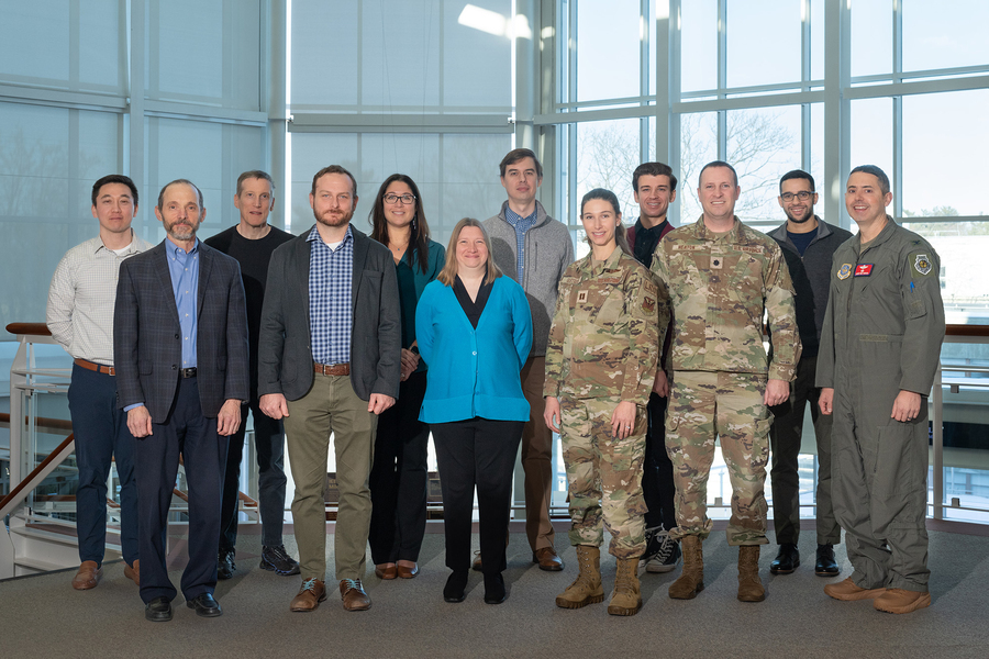 A group of a dozen people, including several in military camouflage uniforms and one in a flight suit, pose together in a bright atrium with large windows.