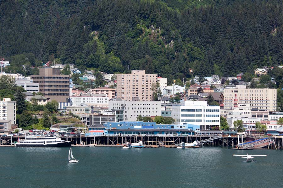 Juneau, Alaska waterfront with seaplanes docked at the pier, a sailboat on the channel, and the city climbing a forested hillside.