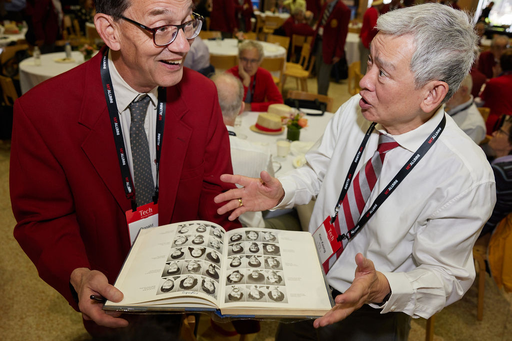 Two men review an open yearbook containing a grid of black and white photographs at an indoor event.