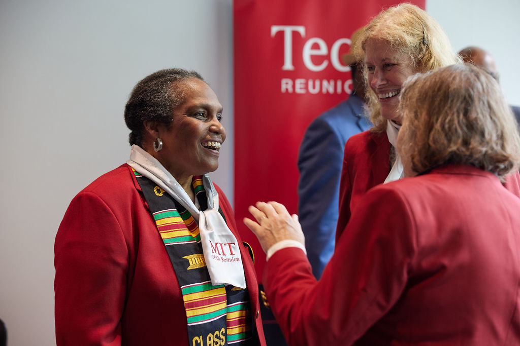 Three women conversing at Tech Reunions event, one wearing red blazer with colorful striped scarf.