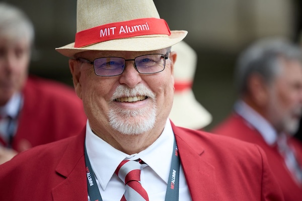 Older man wearing red MIT Alumni blazer, glasses, and straw hat with red band, smiling outdoors.