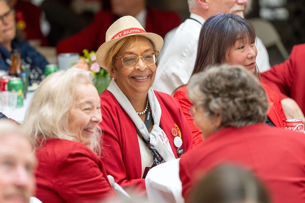 Woman in red blazer and MIT alumni hat smiling among other attendees seated at a dining table.