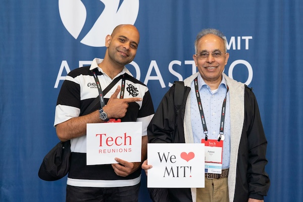Two men standing before blue MIT backdrop, holding signs reading "Tech REUNIONS" and "We ❤ MIT!”