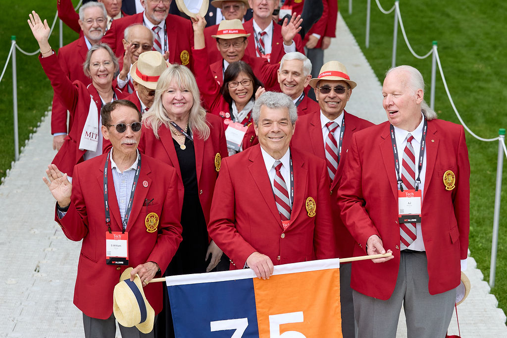 Group of MIT alumni wearing red blazers and holding a banner marked "75" on a grassy pathway.
