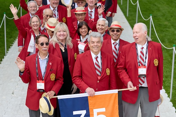 Group of MIT alumni wearing red blazers and holding a banner marked "75" on a grassy pathway.