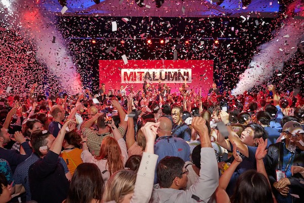 Large indoor celebration event with crowd and falling confetti beneath "MITALUMNI" signage and purple and red lighting.