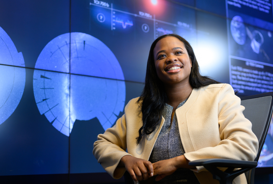 MIT researcher smiling in front of large display screens showing astronomical data and radar imagery in a space science visualization lab.