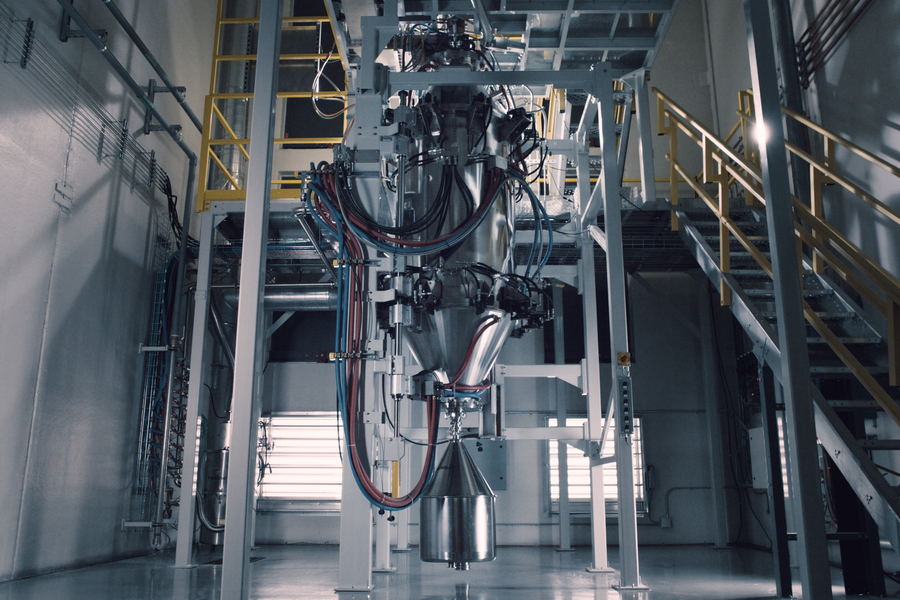 A large industrial processing machine with a polished metal hopper and colorful tubing suspended within a steel-framed facility.
