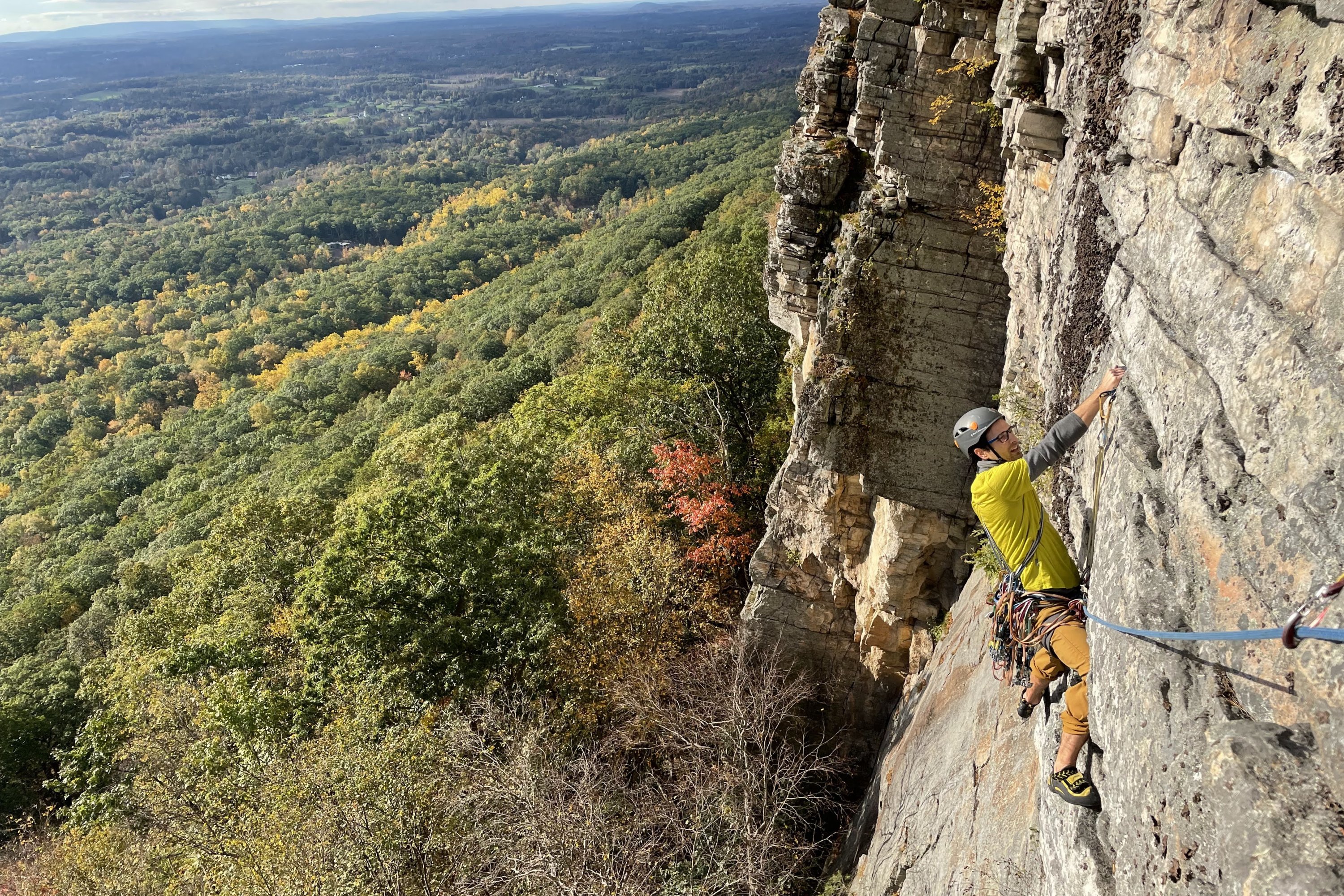 A rock climber in a yellow shirt and helmet traverses a steep cliff face above a vast expanse of New England forest in peak autumn foliage.