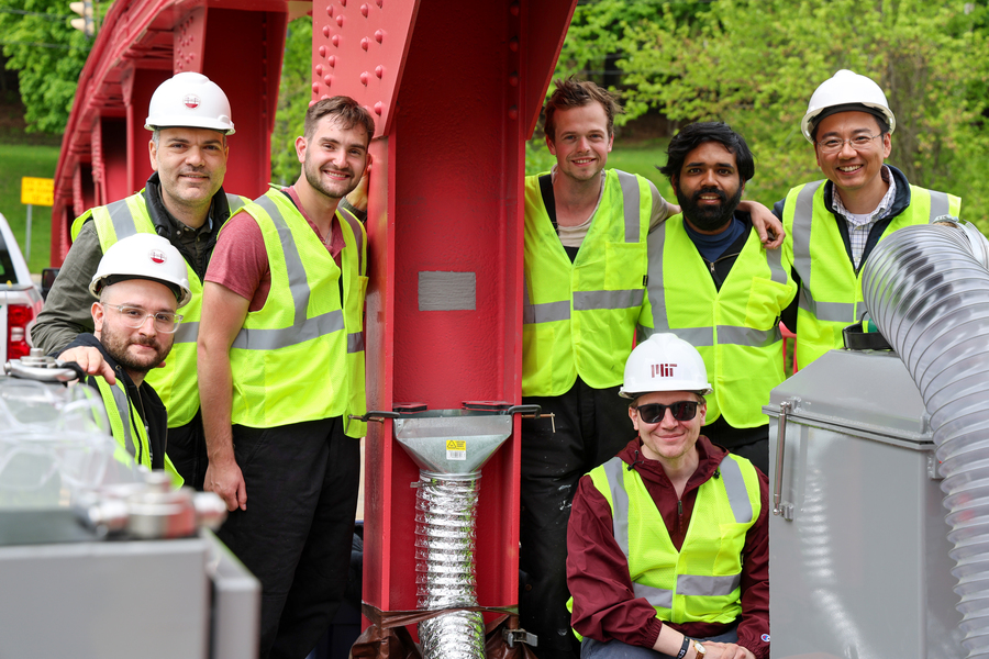 Seven researchers in yellow safety vests and hard hats pose together around industrial equipment attached to a red steel structure outdoors.