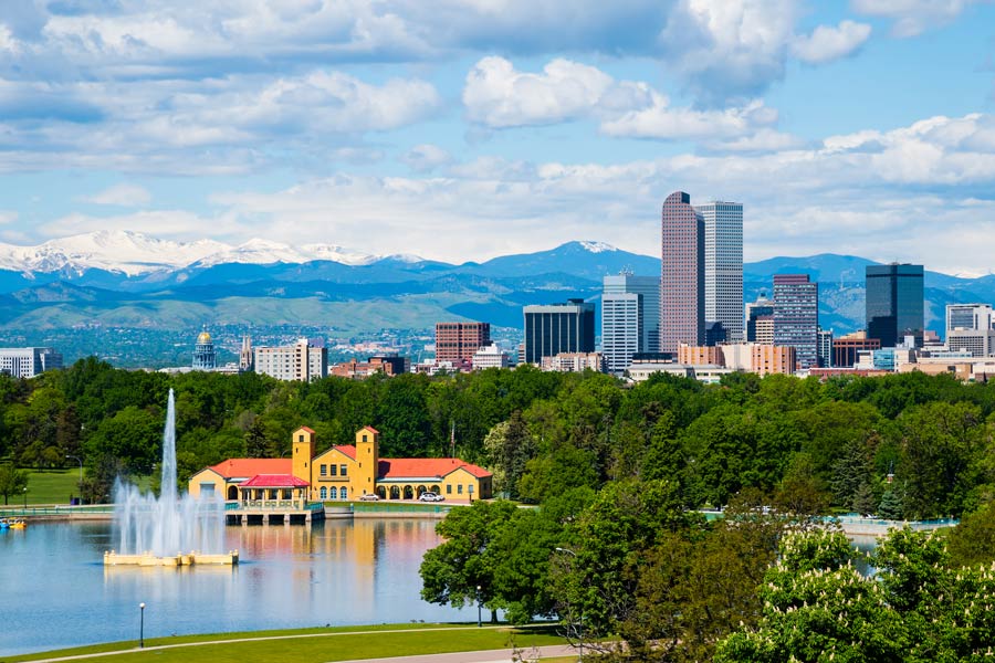 Denver skyline with City Park boathouse and fountain in the foreground; Rocky Mountains with snow-capped peaks in the background.