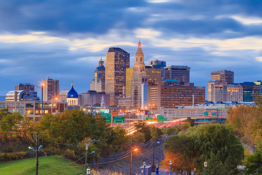 Hartford, Connecticut skyline at dusk with highway light trails, historic church domes, and the Travelers Tower rising above the city.