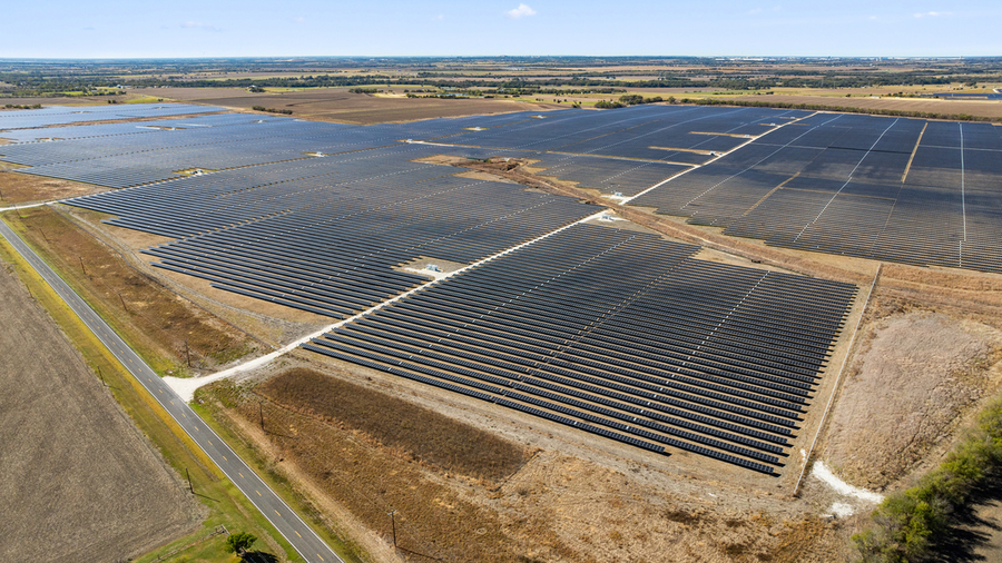 Aerial view of a large utility-scale solar farm with rows of photovoltaic panels stretching across flat agricultural land under a partly cloudy sky