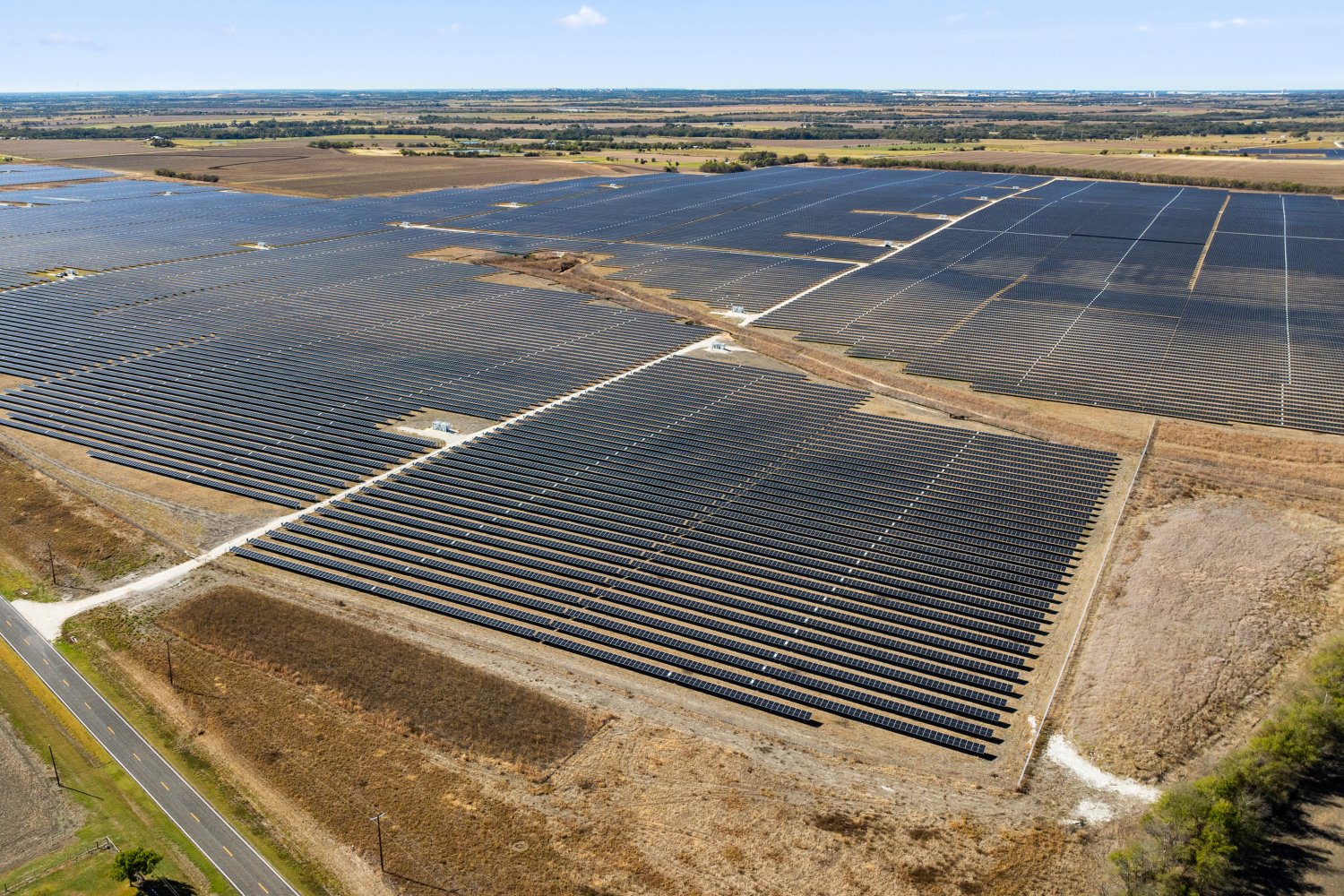 Aerial view of a large utility-scale solar farm with rows of photovoltaic panels stretching across flat agricultural land under a partly cloudy sky