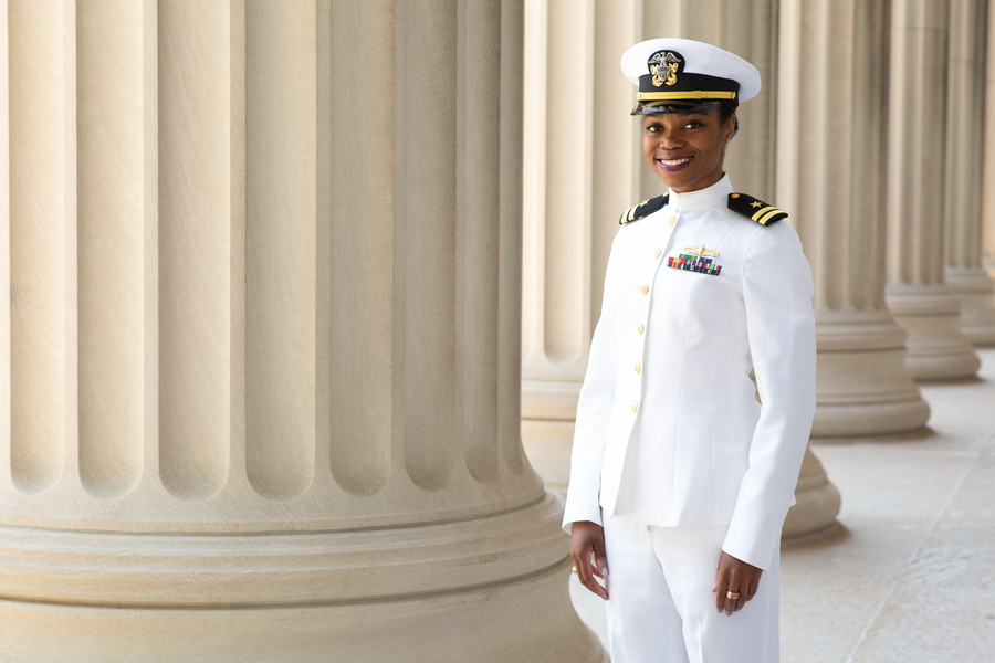 A smiling Naval officer in full white dress uniform with ribbons and insignia stands between stone columns on a classical campus colonnade.