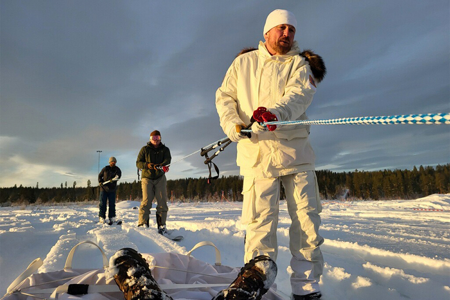Three people in winter gear manage a blue and white tow rope on a snow-covered open field at low sun, with a pine forest in the background.