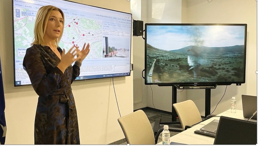 Female presenter gesturing at dual screens displaying geographic maps with location markers and a landscape view with geothermal features during a research presentation.