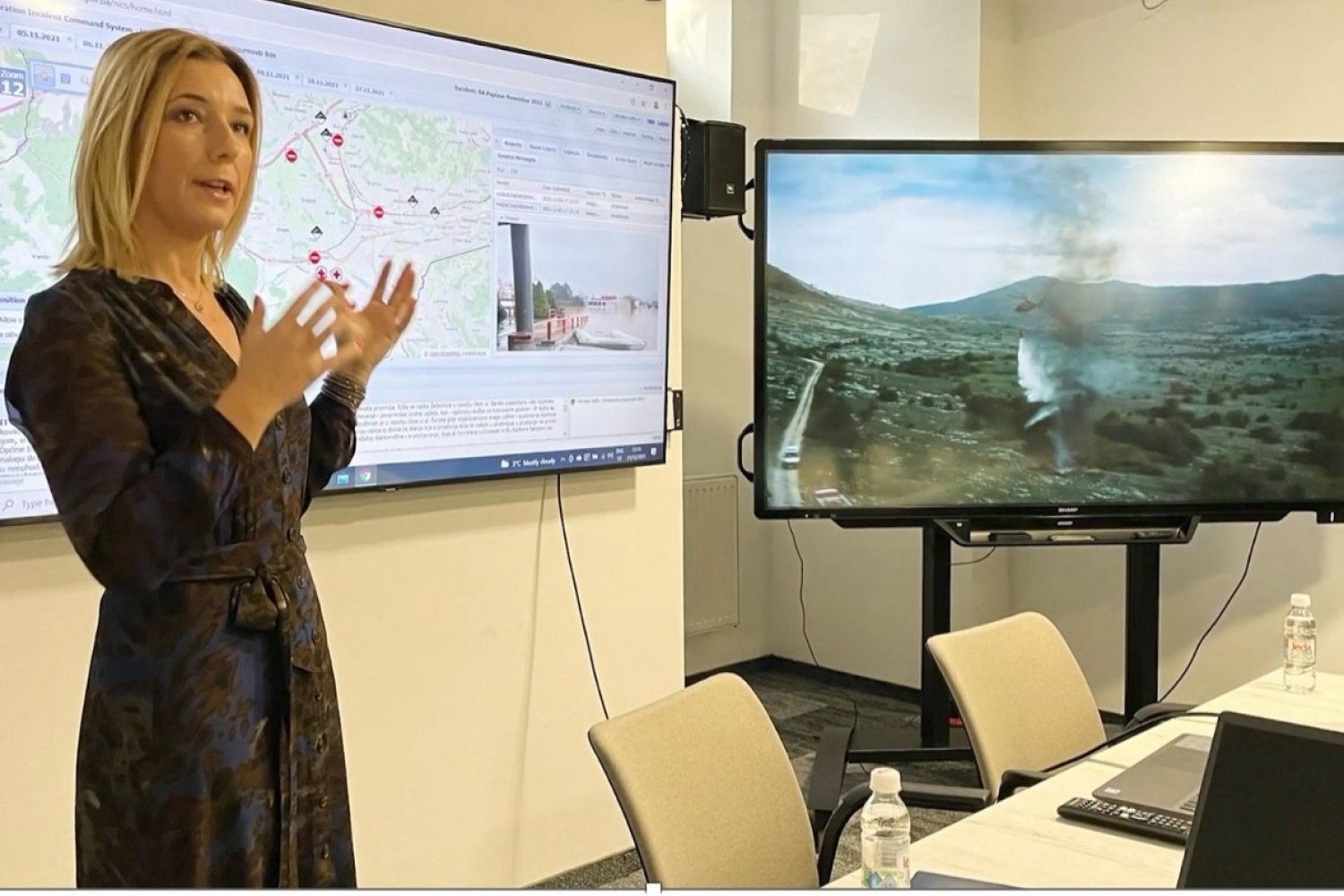 Female presenter gesturing at dual screens displaying geographic maps with location markers and a landscape view with geothermal features during a research presentation.