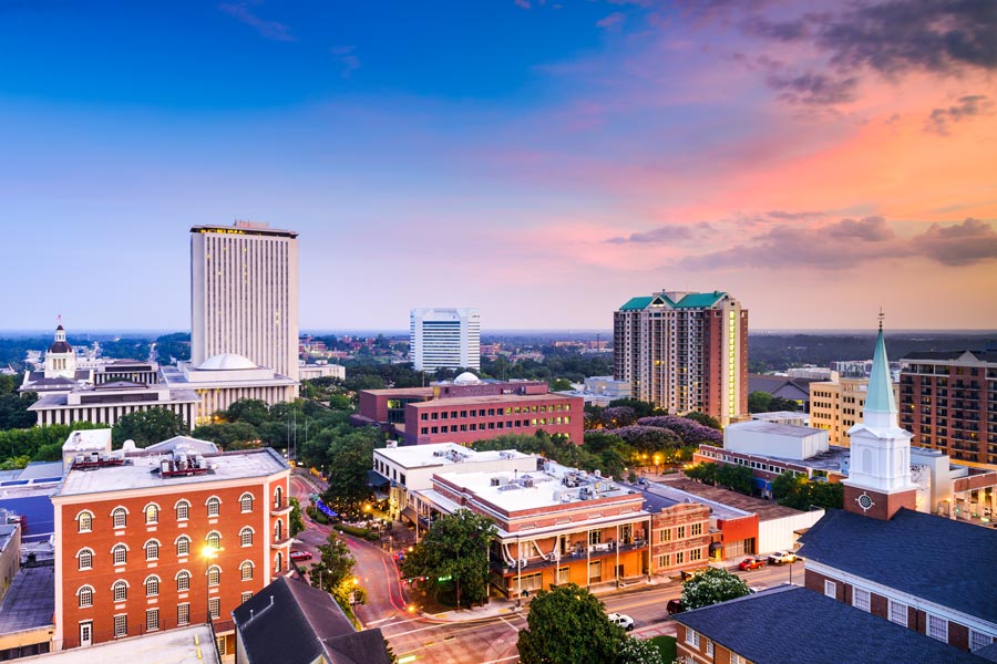 Tallahassee, Florida skyline at sunset, with the Florida State Capitol building visible at left and a white church steeple amid downtown streets and summer foliage.