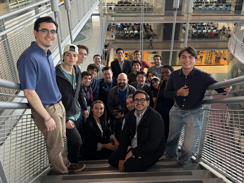 A large group of smiling students and professionals posing together on a staircase inside a modern, multi-level academic building.
