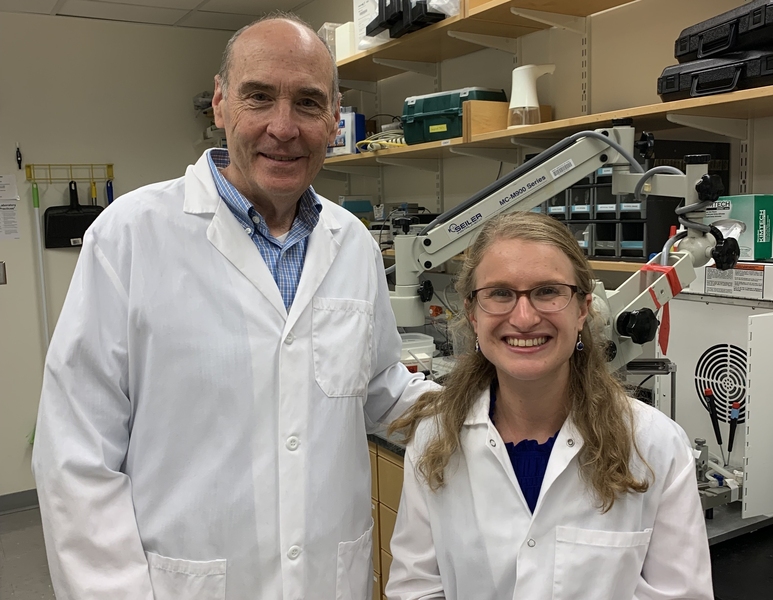 Two researchers in white lab coats smile at the camera in a laboratory equipped with a microscope and scientific instruments.