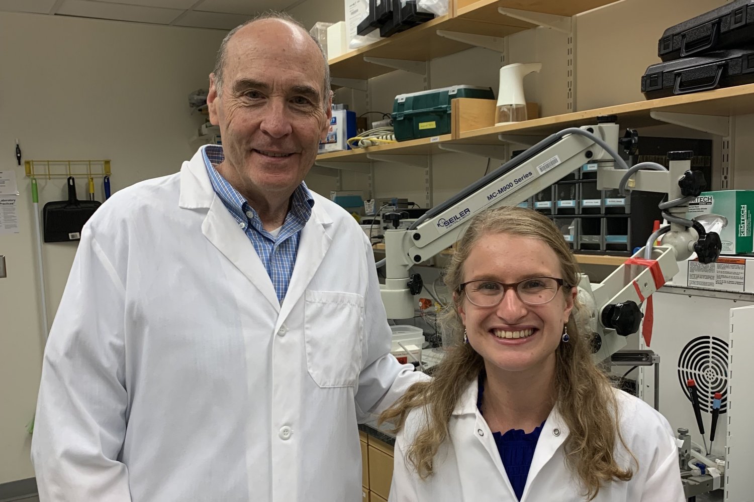 Two researchers in white lab coats smile at the camera in a laboratory equipped with a microscope and scientific instruments.