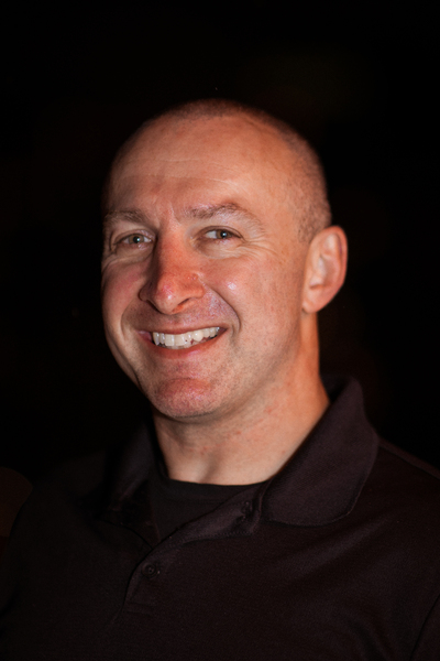 A person smiling against a dark background, wearing a black polo shirt.