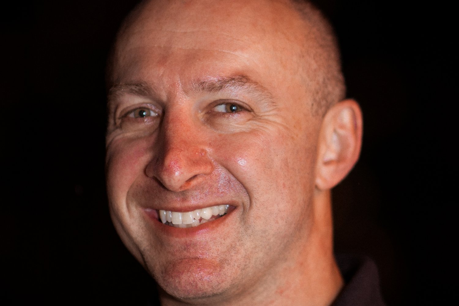 A person smiling against a dark background, wearing a black polo shirt.