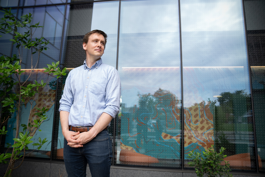 A man in a light blue shirt stands with hands clasped in front of a large glass building facade reflecting trees and a colorful interior mural.