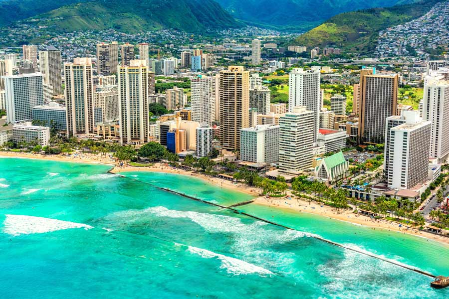 Aerial view of Waikiki Beach in Honolulu, Hawaii, with turquoise ocean waters, palm-lined shore, and high-rise hotels backed by the Ko'olau Mountains.