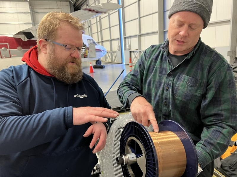 Two technicians examine a large circular component with blue casing and copper elements in an industrial warehouse setting.