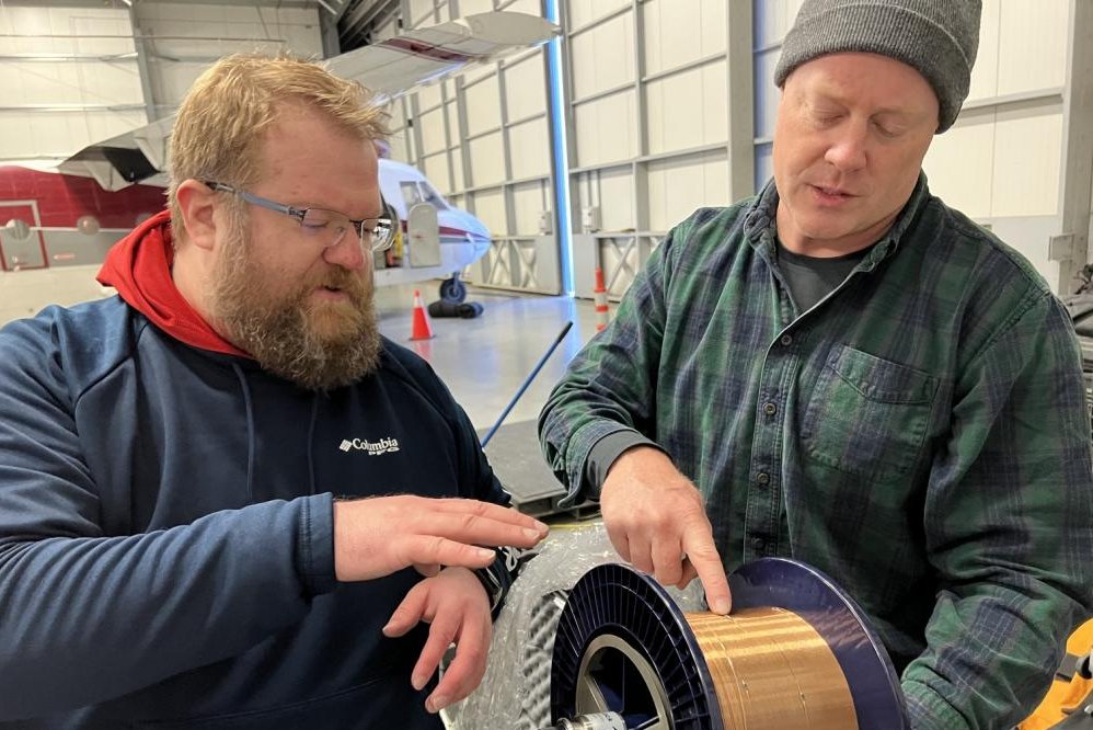Two technicians examine a large circular component with blue casing and copper elements in an industrial warehouse setting.