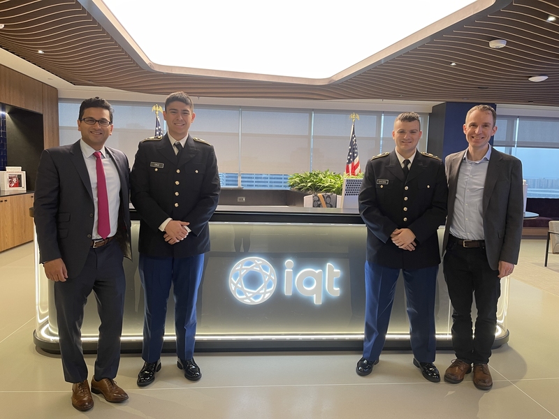 Five men stand before illuminated IQT logo in modern office: two in military uniforms, three in business suits, with cityscape visible through windows.