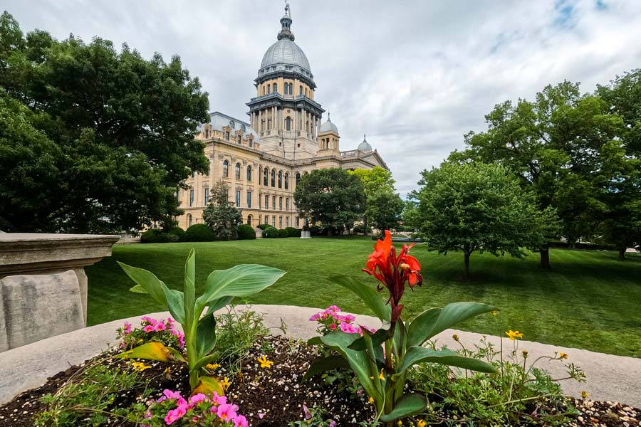 The Illinois State Capitol building in Springfield, viewed from a summer flower garden, with its large silver dome rising above ornate stone facades.