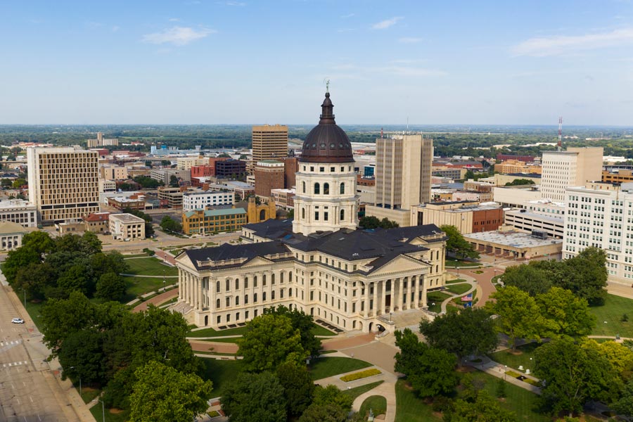 Aerial view of the Kansas State Capitol in Topeka, featuring its large copper dome topped with a bronze statue, surrounded by manicured grounds and the downtown skyline.