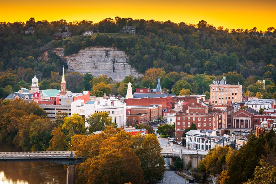 Aerial view of downtown Frankfort, Kentucky at sunset, with church steeples, brick buildings, fall foliage, and a limestone bluff rising behind the city.