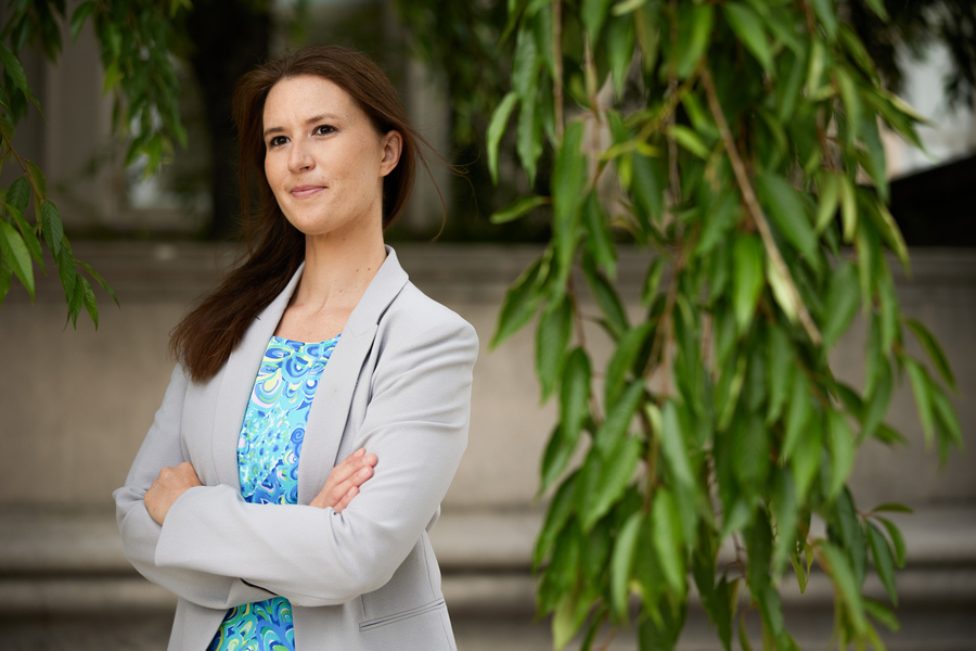 A woman in a light gray blazer stands with arms crossed outdoors, with hanging willow branches in the background.