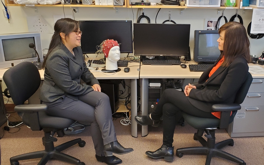 Diane Chan and Li-Huei Tsai sit, talking, at a table covered in research equipment. In front of powered-off monitors sits a polystyrene head with a red cap of electrodes.