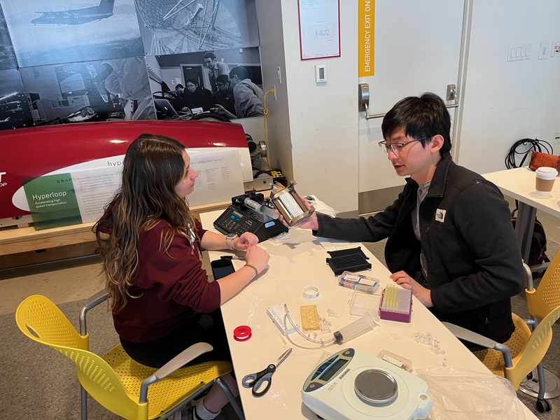 Two researchers collaborate at a worktable covered with lab equipment and components, with a red Hyperloop pod model and historical photographs visible in the background.