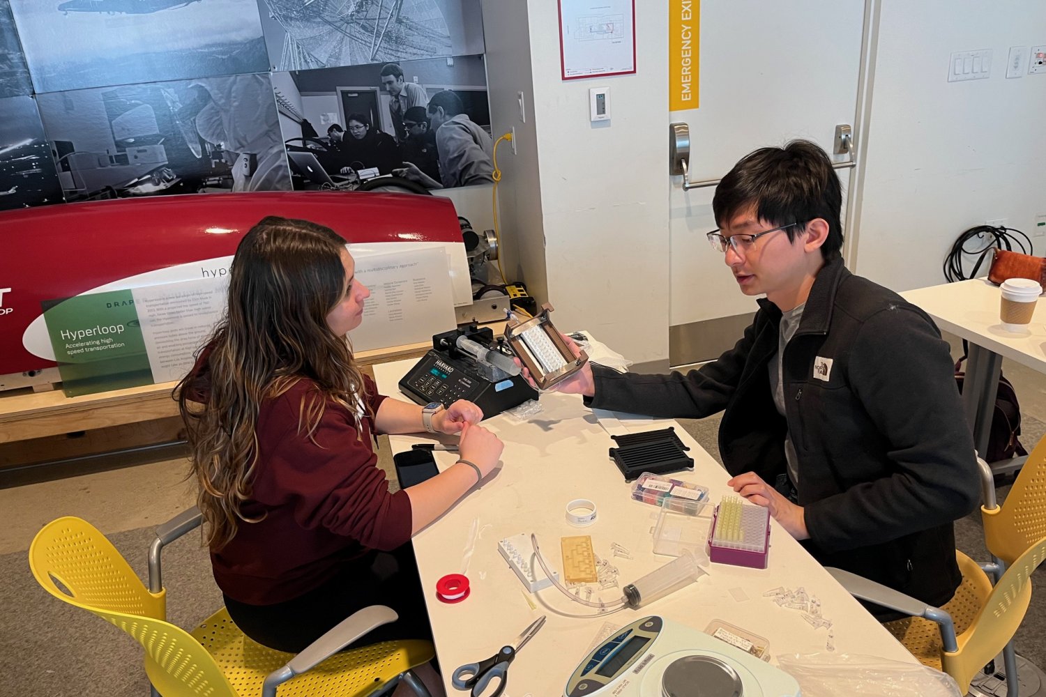 Two researchers collaborate at a worktable covered with lab equipment and components, with a red Hyperloop pod model and historical photographs visible in the background.