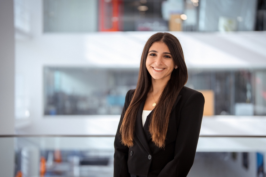 A smiling young woman in a black blazer stands in a modern atrium with glass railings and an open multi-story workspace behind her.
