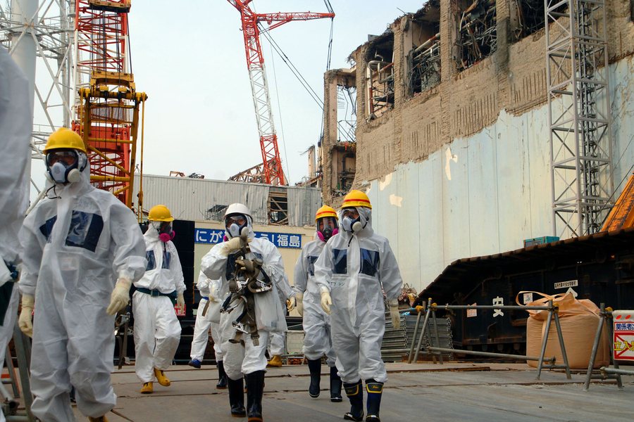 Workers in white protective suits, respirators, and hard hats walk past the damaged exterior of the Fukushima Daiichi nuclear power plant during decommissioning operations.