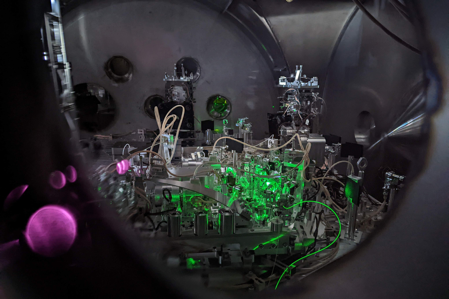 Interior view through a circular viewport into a LIGO interferometer vacuum chamber, revealing densely packed optical components and mirrors lit by vivid green laser beams with a pink laser visible at the edge.