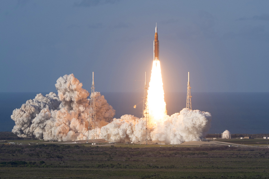 A large rocket lifts off from a launch pad in a burst of flame and billowing smoke against a cloudy sky.