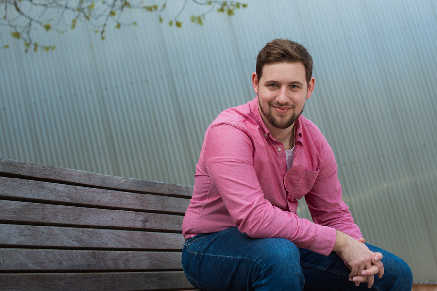 Man in pink button-up shirt and blue jeans sitting on wooden bench against corrugated metal wall.
