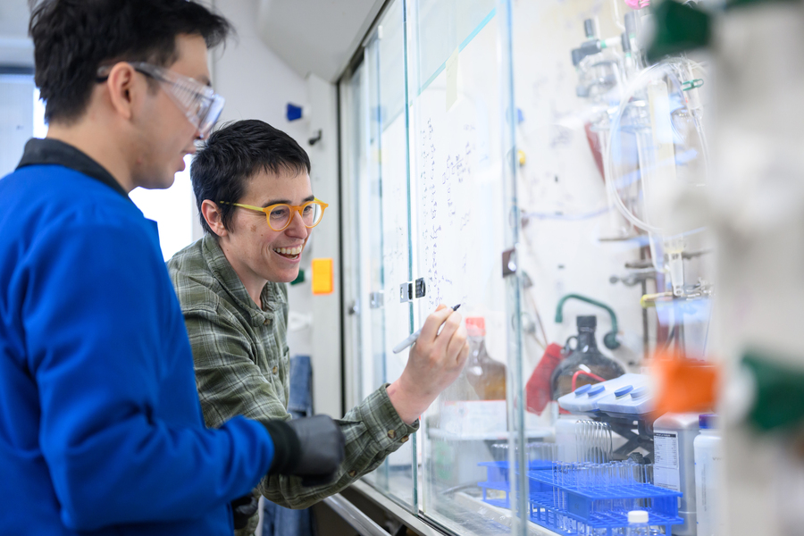 A person smiling and writing chemical structures on a glass surface in a laboratory, alongside a colleague in a blue lab coat.